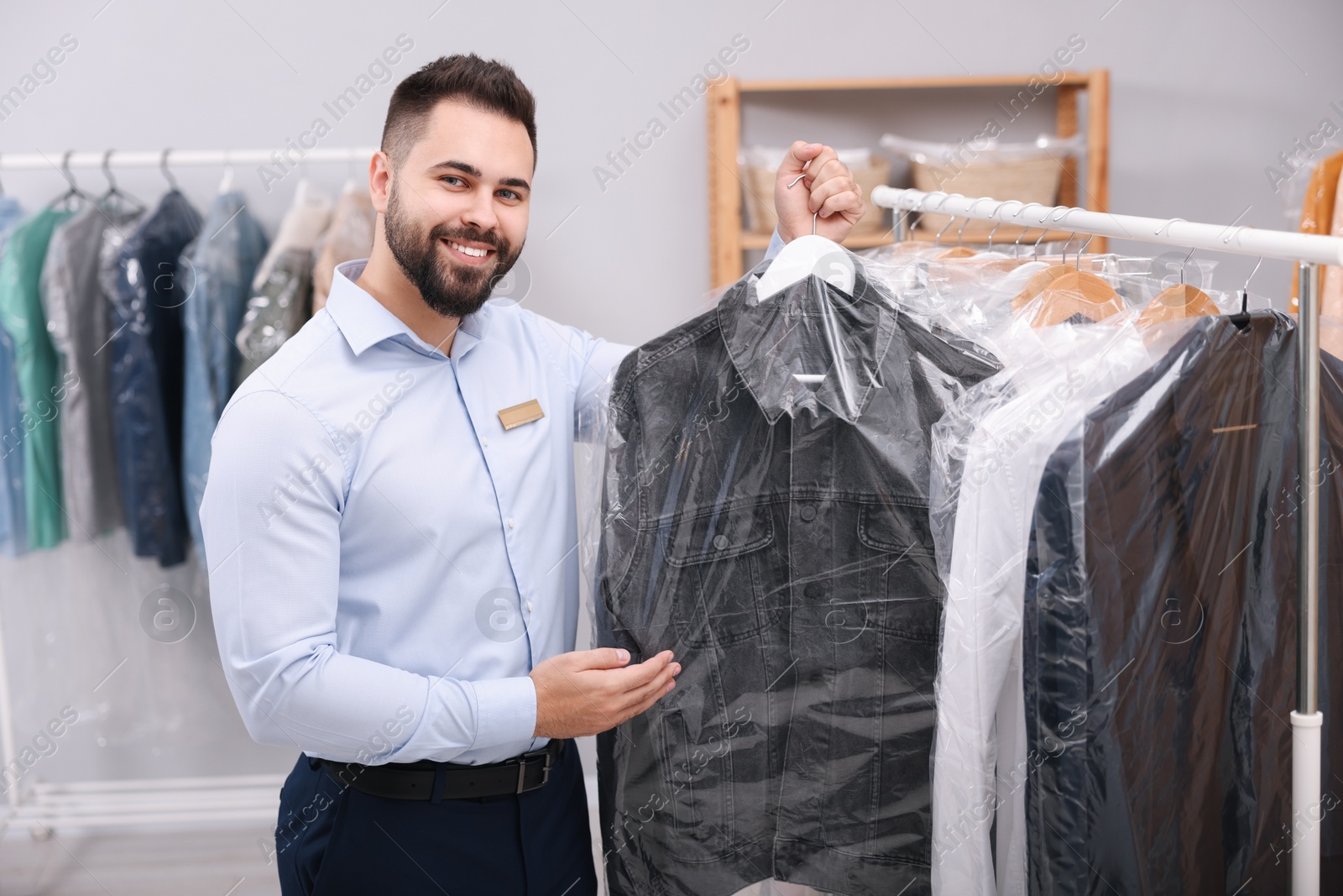 Photo of Dry-cleaning service. Happy worker holding hanger with denim jacket in plastic bag near rack with clothes indoors