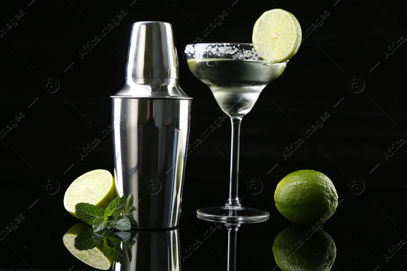 Photo of Metal shaker, delicious cocktail, limes and mint on black mirror surface