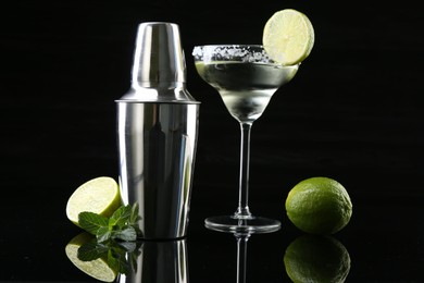 Photo of Metal shaker, delicious cocktail, limes and mint on black mirror surface