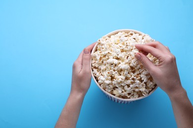 Photo of Woman taking delicious popcorn from paper bucket on light blue background, top view. Space for text