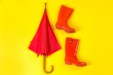 Photo of Beautiful red umbrella and rubber boots on light yellow background, flat lay