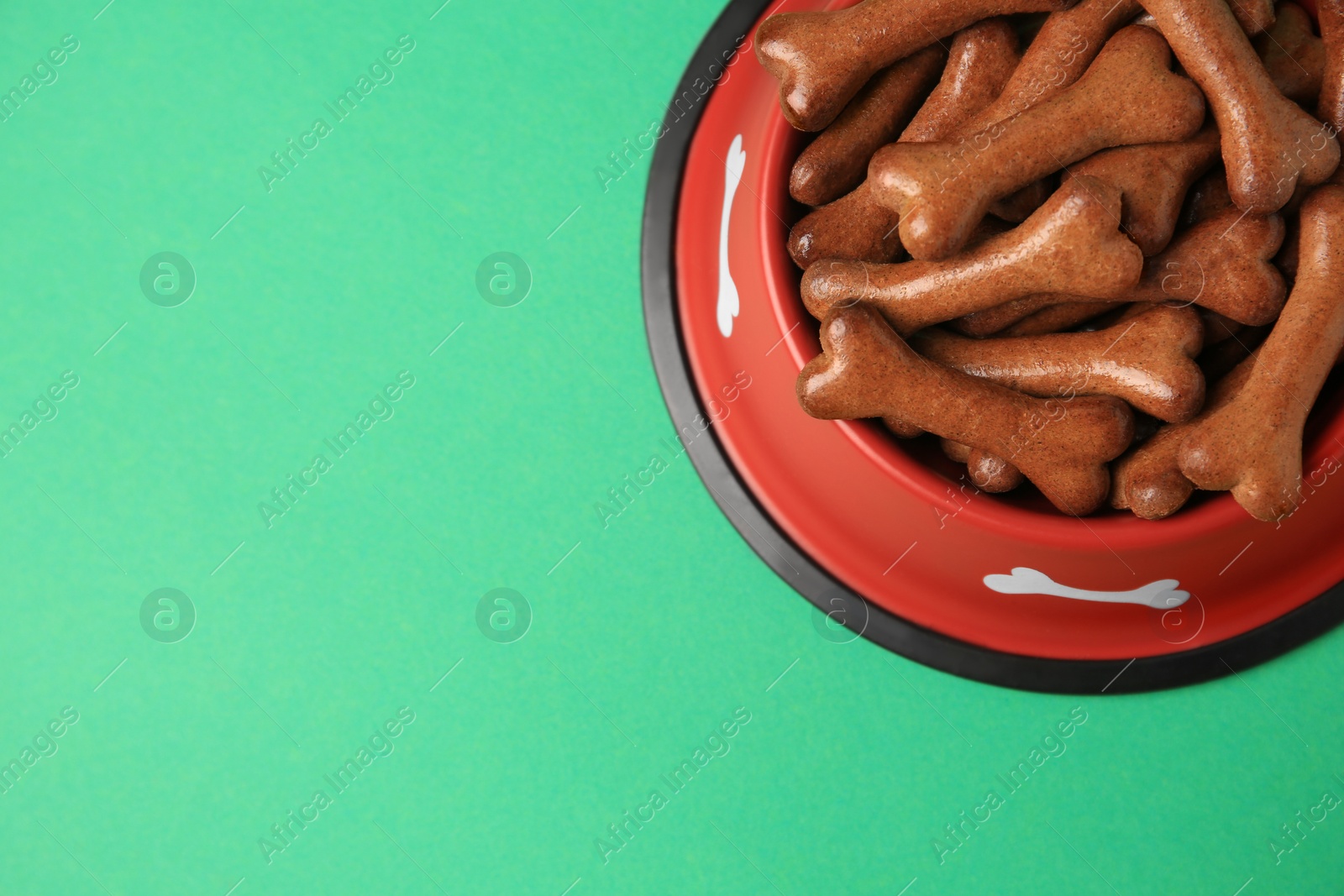 Photo of Red bowl with bone shaped dog cookies on green background, top view. Space for text