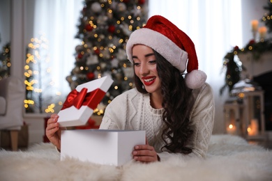 Photo of Young woman wearing Santa hat opening Christmas gift on floor at home