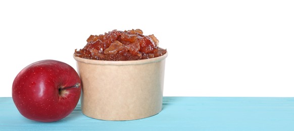 Delicious apple jam in paper cup and fresh fruit on light blue wooden table against white background, space for text