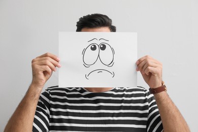 Man hiding emotions using card with drawn frowning face on white background