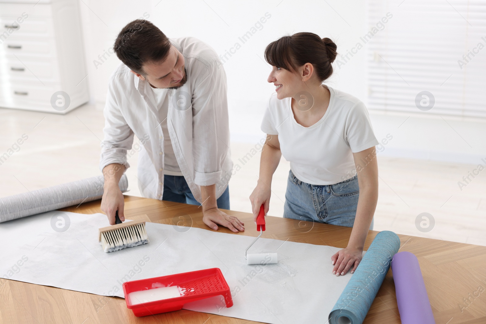 Photo of Woman and man applying glue onto wallpaper sheet at wooden table indoors