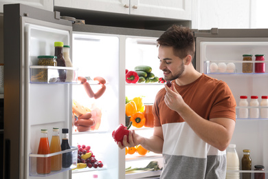 Photo of Young man with bell pepper and grape berry near open refrigerator indoors