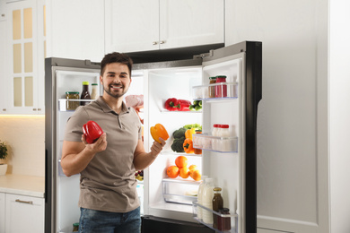 Photo of Young man with bell peppers near open refrigerator indoors