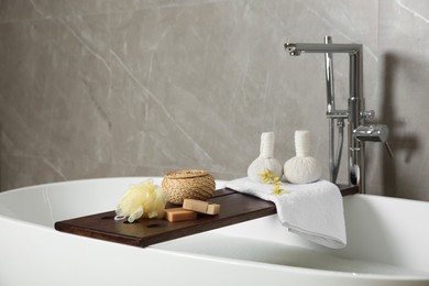 Photo of Wooden bath tray with herbal massage bags and bathroom amenities on tub indoors