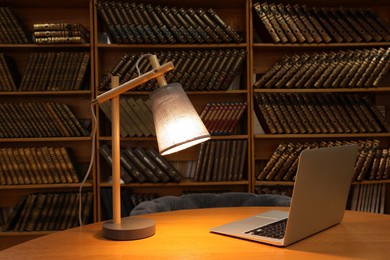 Photo of Lamp and laptop on wooden table near shelves with collection of vintage books in library