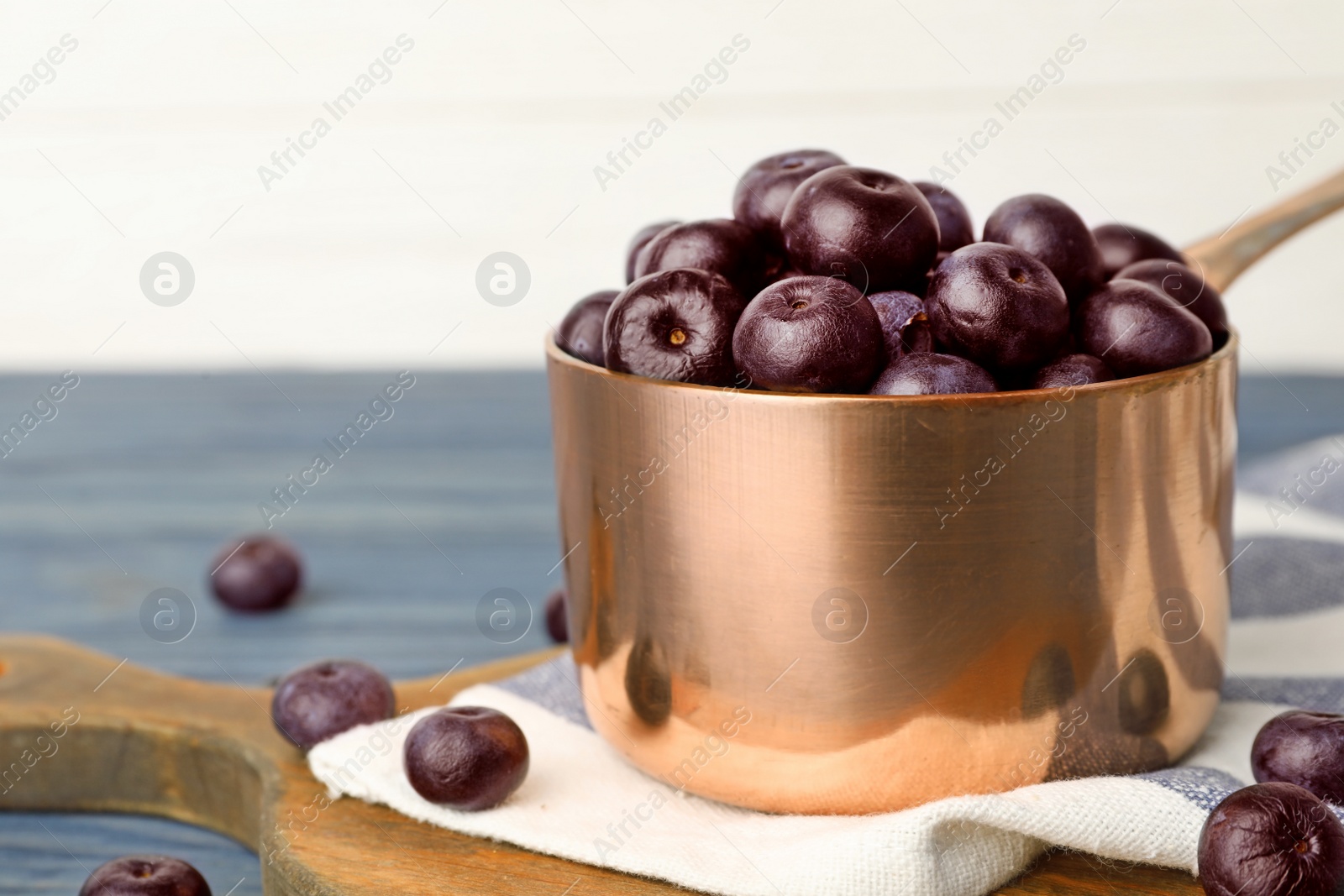 Photo of Metal saucepan of fresh acai berries on blue wooden table against white background, closeup. Space for text