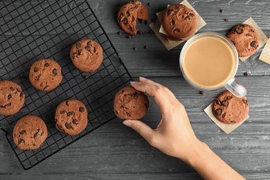 Photo of Woman taking tasty chocolate chip cookie from cooling rack on wooden background, top view
