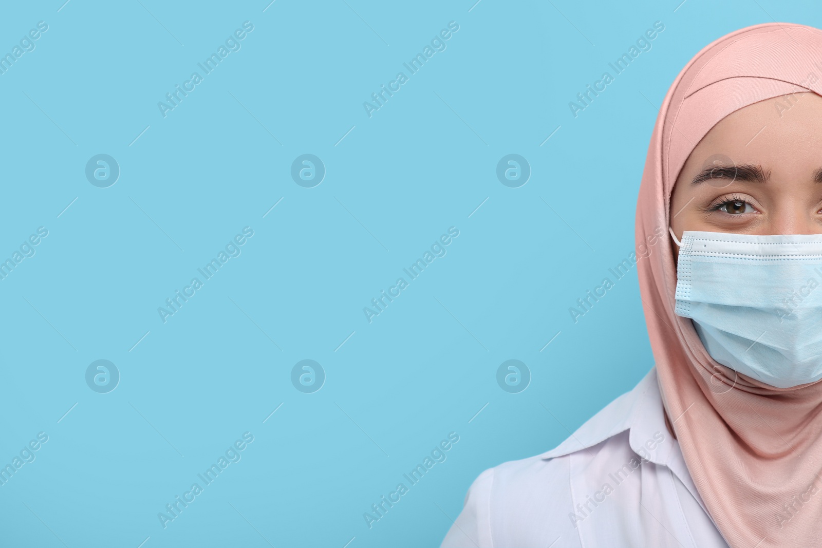 Photo of Muslim woman wearing hijab, medical uniform and protective mask on light blue background, closeup. Space for text