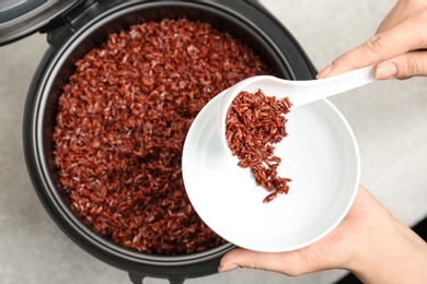 Woman putting brown rice into bowl from multi cooker on grey background, top view