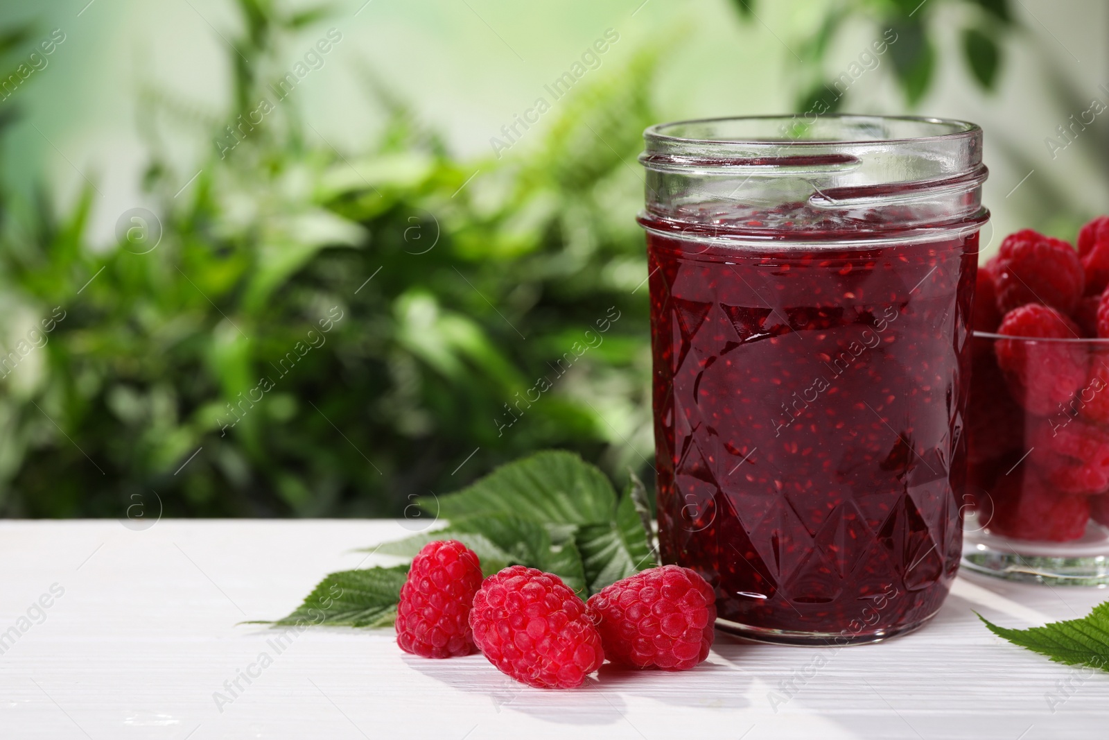 Photo of Delicious jam in glass jar and fresh raspberries on white wooden table. Space for text