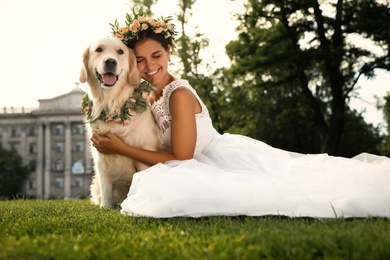 Bride and adorable Golden Retriever wearing wreath made of beautiful flowers on green grass outdoors