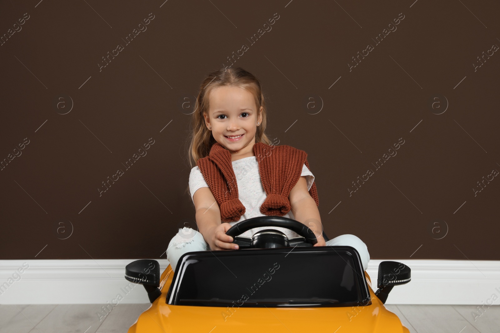 Photo of Cute little girl driving children's electric toy car near brown wall indoors