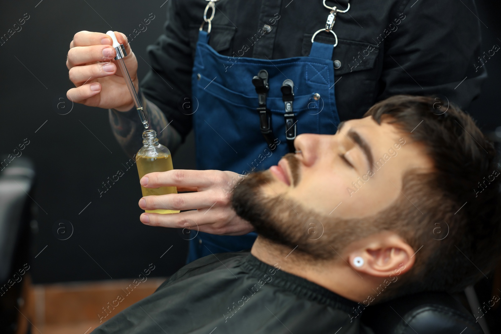 Photo of Hairdresser with beard oil near client in barbershop, closeup. Professional shaving service