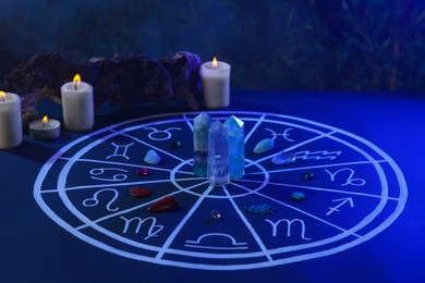 Photo of Natural stones for zodiac signs, drawn astrology chart and burning candles on dark blue table
