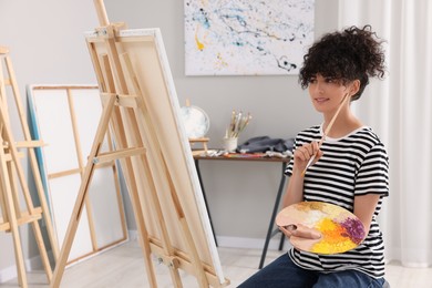 Photo of Young woman holding brush and artist`s palette near easel with canvas in studio