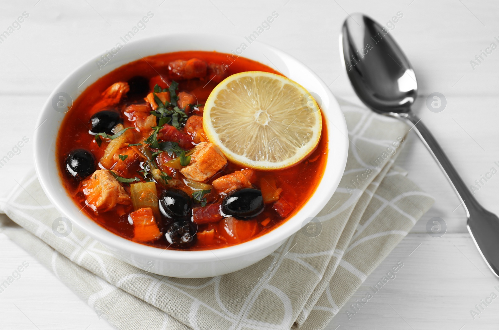 Photo of Meat solyanka soup with sausages, olives, vegetables and spoon on white wooden table