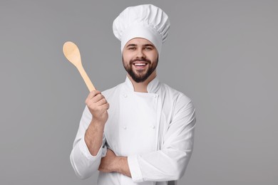 Photo of Happy young chef in uniform holding wooden spoon on grey background