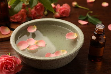Photo of Bowl of water, bottle with essential oil and beautiful rose petals on wooden table. Aromatherapy treatment