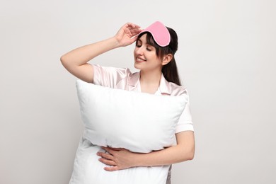 Happy woman in pyjama and sleep mask holding pillow on light grey background