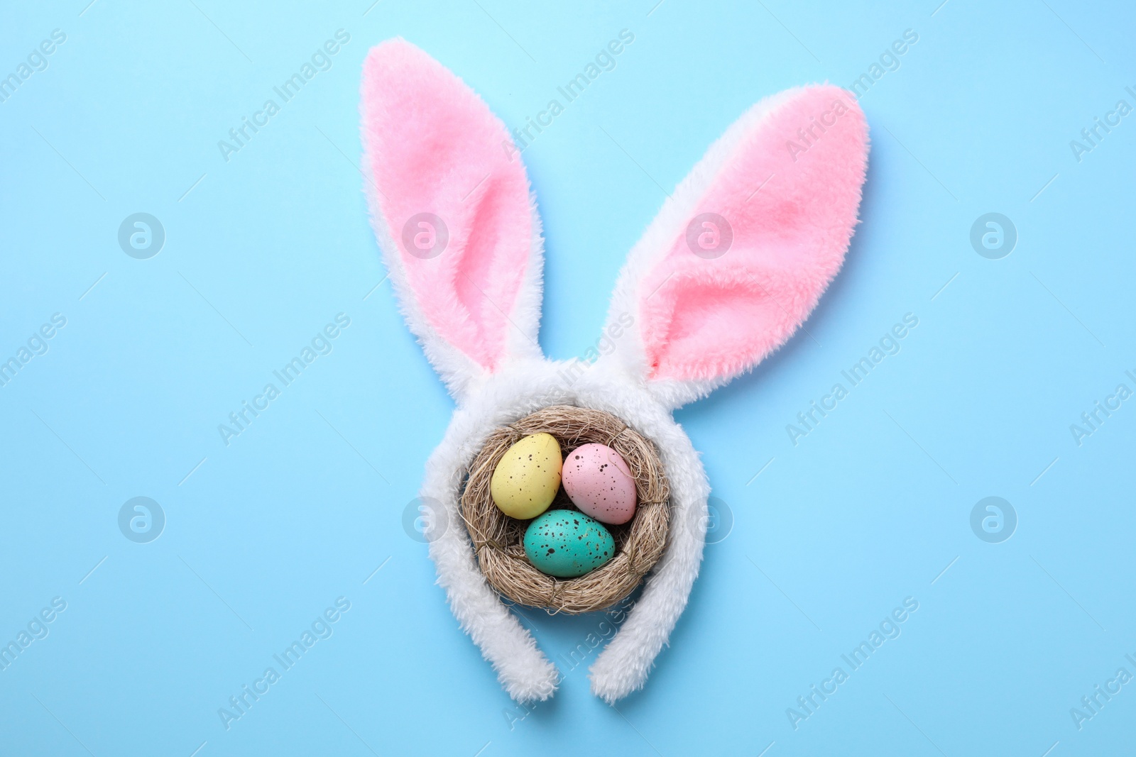 Photo of Headband with Easter bunny ears and dyed eggs in nest on light blue background, flat lay