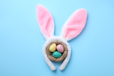 Headband with Easter bunny ears and dyed eggs in nest on light blue background, flat lay