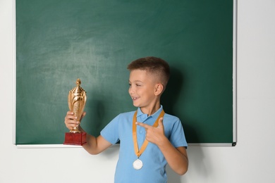 Photo of Happy boy with golden winning cup and medal near chalkboard in classroom