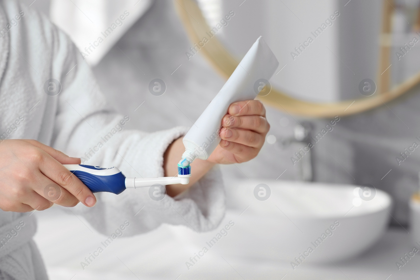 Photo of Woman squeezing toothpaste from tube onto electric toothbrush in bathroom, closeup