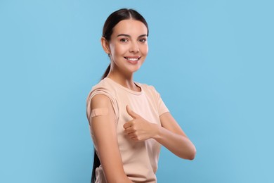 Photo of Woman with sticking plaster on arm after vaccination showing thumbs up against light blue background