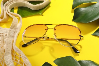 Photo of Stylish sunglasses, net bag and green leaves on yellow background