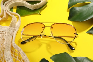 Stylish sunglasses, net bag and green leaves on yellow background