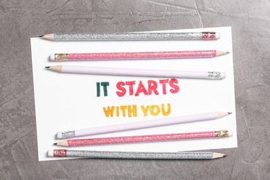 Photo of Sheet of paper with phrase It Starts With You and pencils on grey table, top view