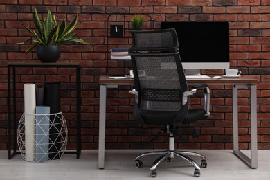 Photo of Cosy workspace with computer on desk, chair and potted plants near brick wall at home