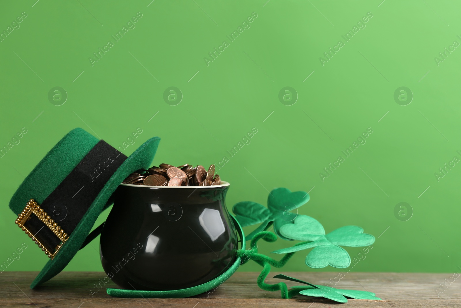 Photo of Pot of gold coins, hat and headband with clover leaves on wooden table against green background, space for text. St. Patrick's Day celebration