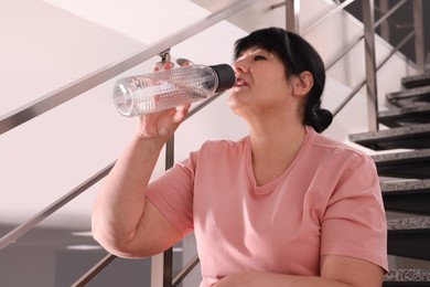 Photo of Overweight mature woman drinking water from bottle on stairs indoors