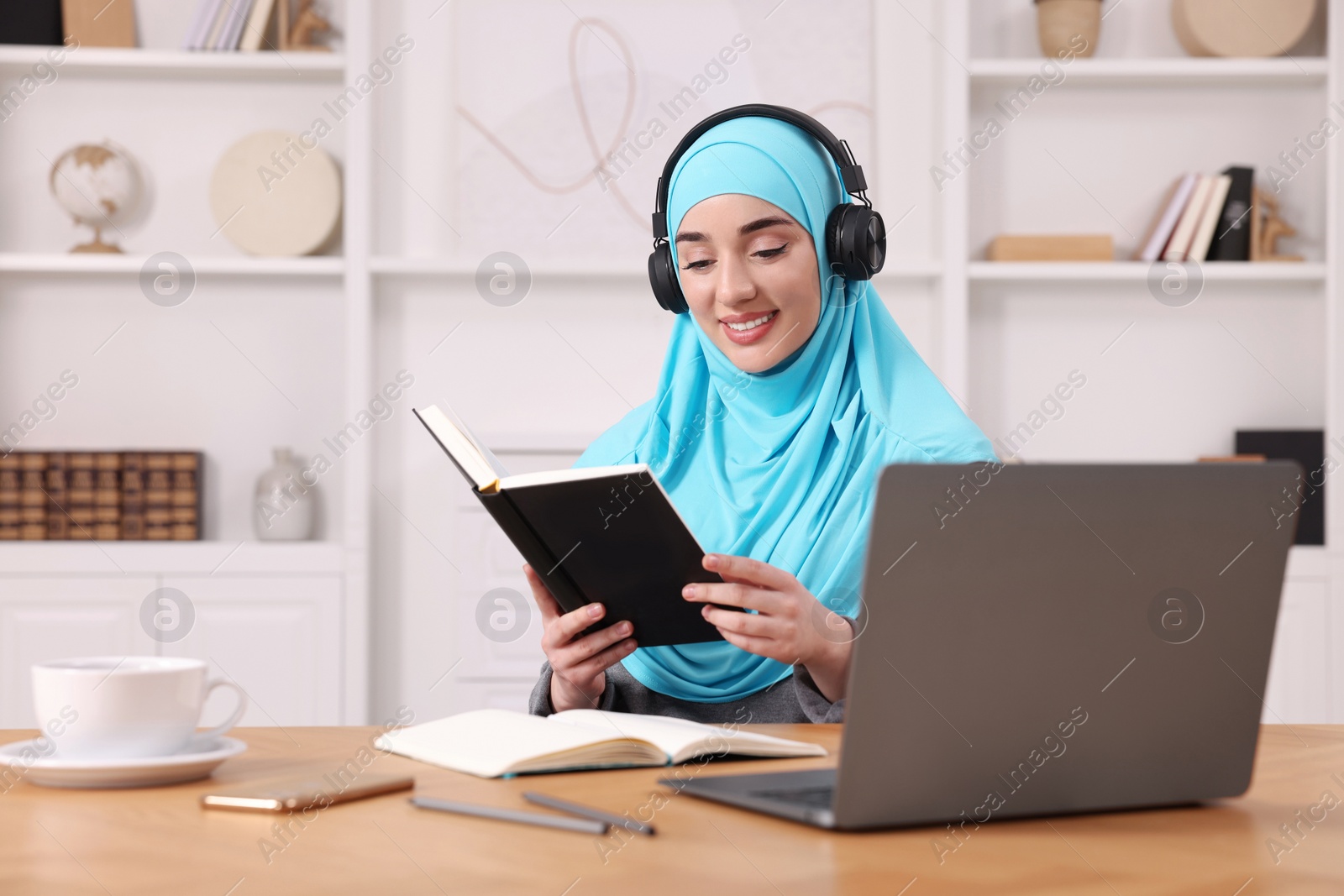 Photo of Muslim woman in headphones studying near laptop at wooden table in room