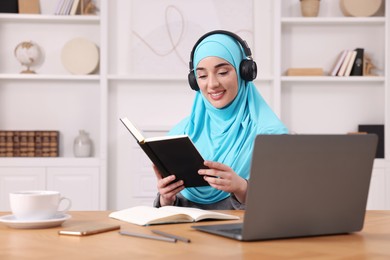 Photo of Muslim woman in headphones studying near laptop at wooden table in room