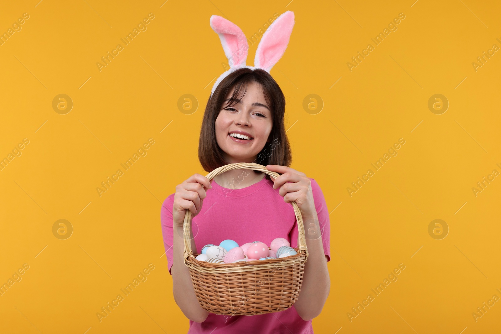 Photo of Easter celebration. Happy woman with bunny ears and wicker basket full of painted eggs on orange background