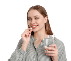 Young woman with glass of water taking vitamin pill on white background