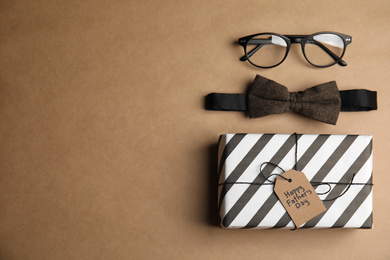 Eyeglasses, bow tie and gift with tag HAPPY FATHER'S DAY on brown background, flat lay. Space for text