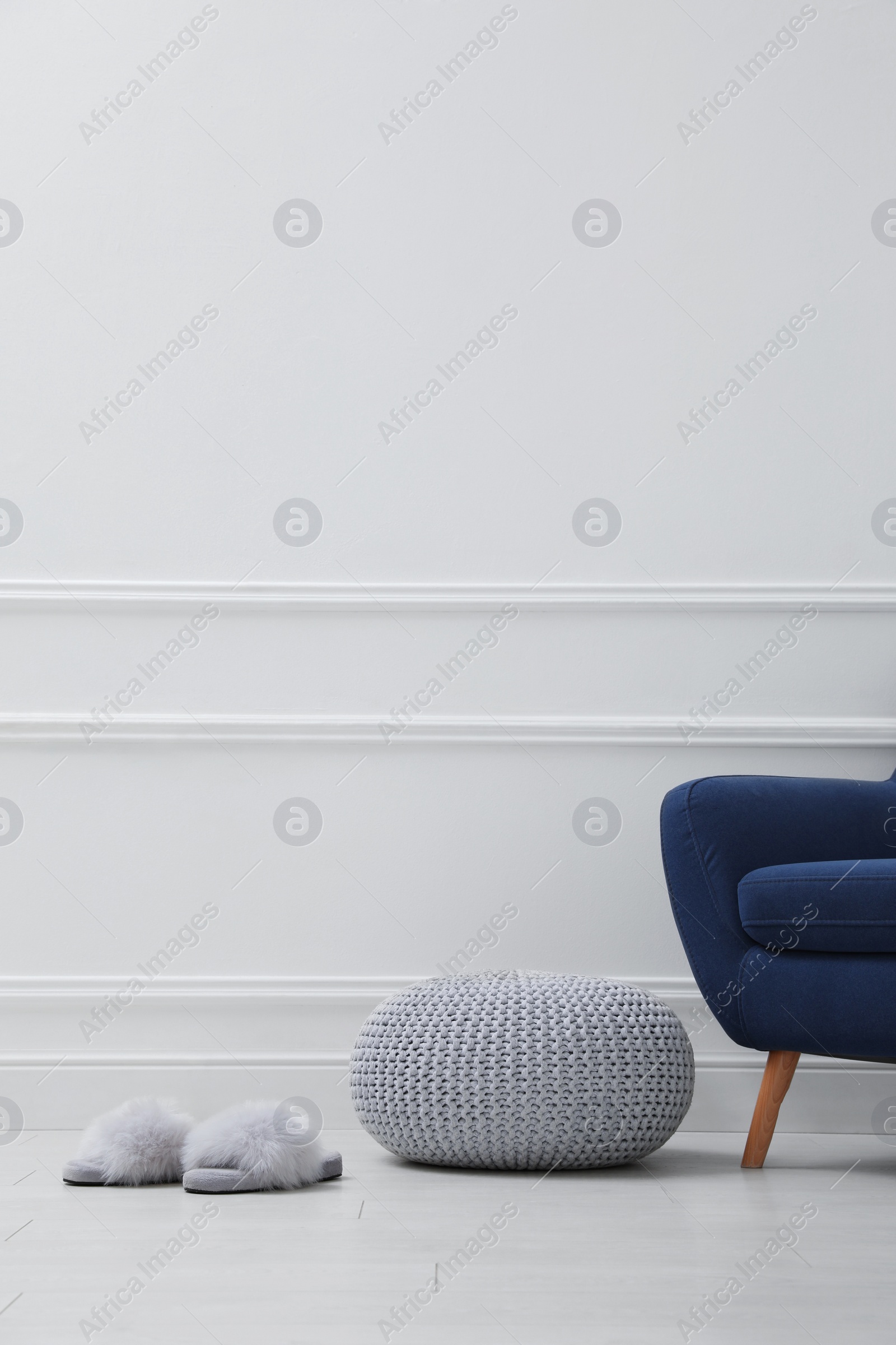 Photo of Knitted pouf, fuzzy slippers and armchair near white wall indoors. Space for text