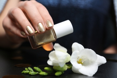 Woman with golden manicure holding nail polish bottle over table, closeup
