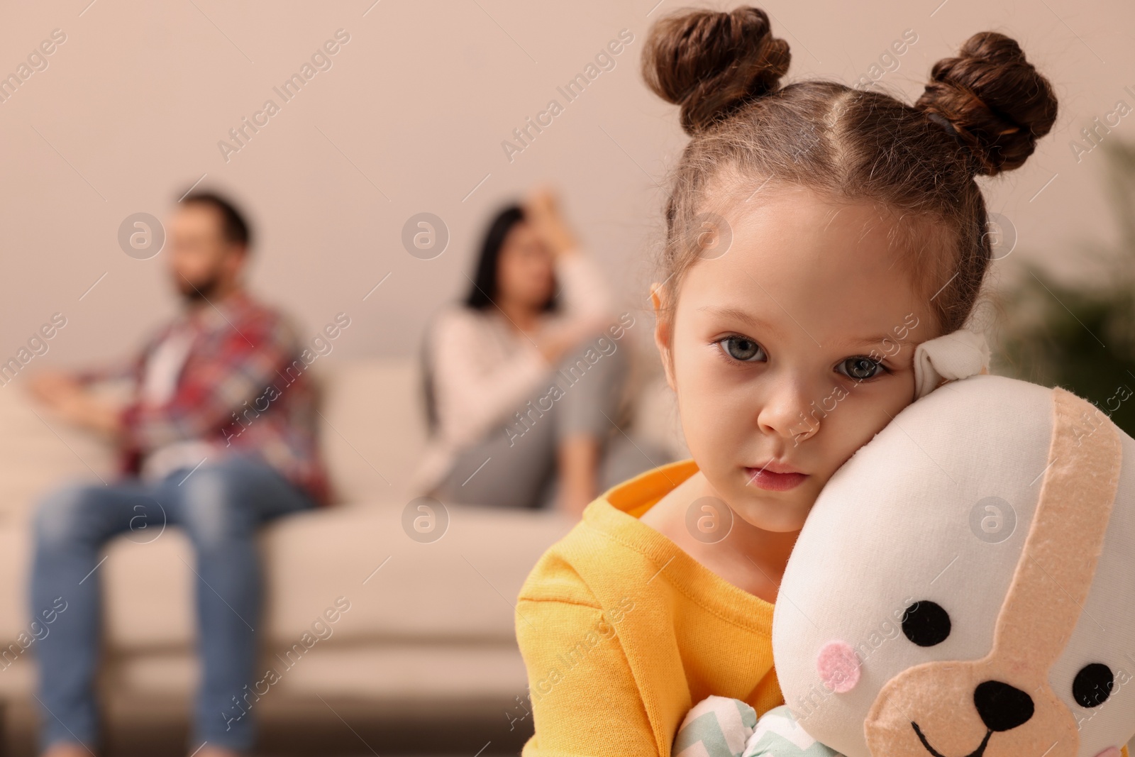 Photo of Sad little girl with toy and her arguing parents on sofa in living room