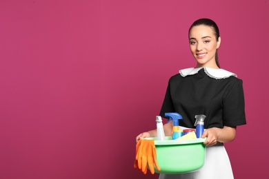 Young chambermaid holding plastic basin with detergents on color background