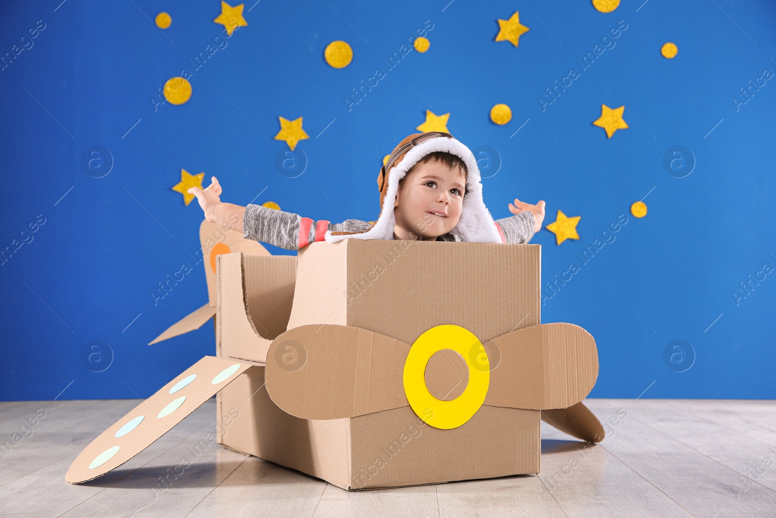 Photo of Cute little child playing with cardboard airplane near blue wall