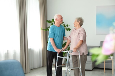 Photo of Elderly man and his wife with walking frame indoors. Space for text